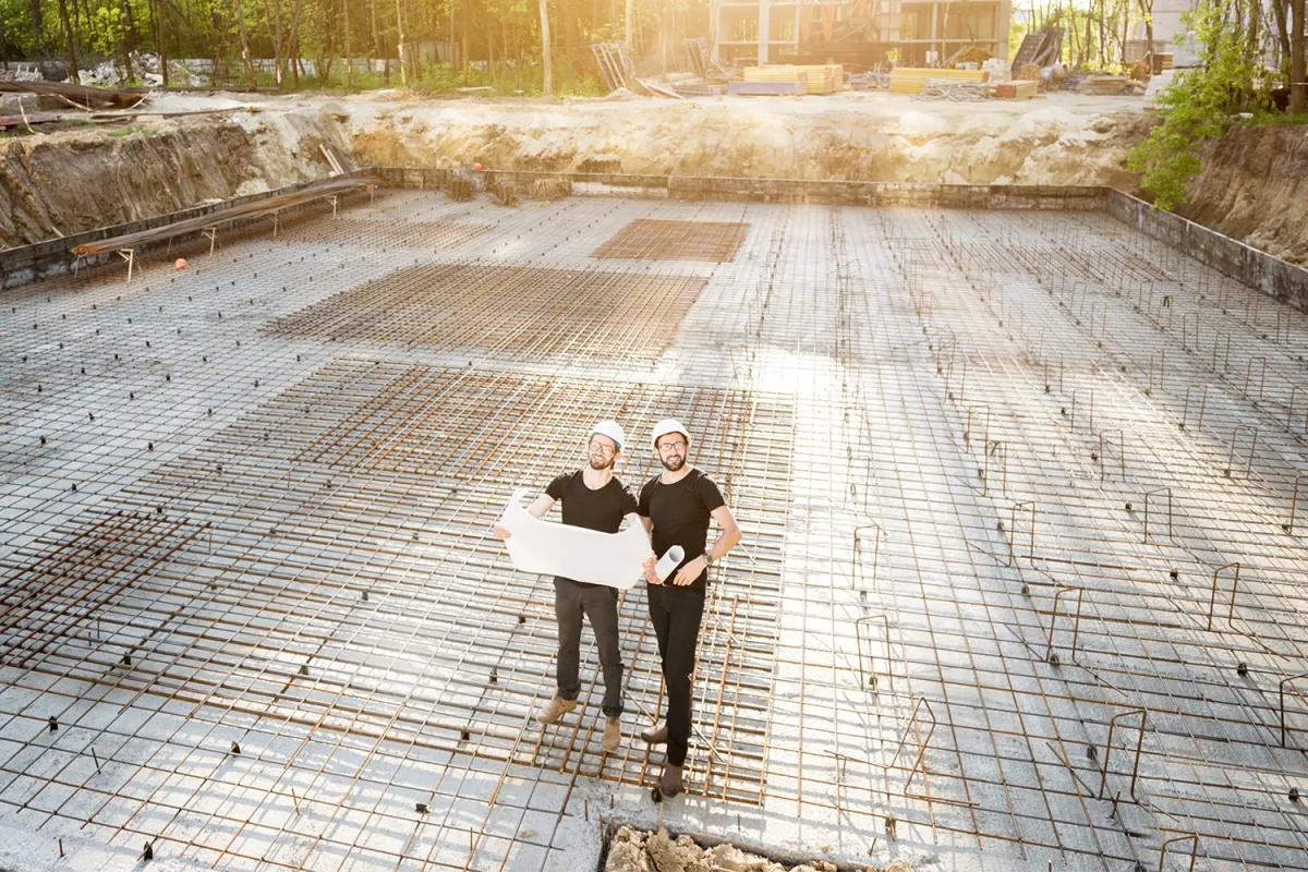 Two construction workers wearing hard hats stand on a reinforced concrete foundation slab, holding blueprints at a building site.
