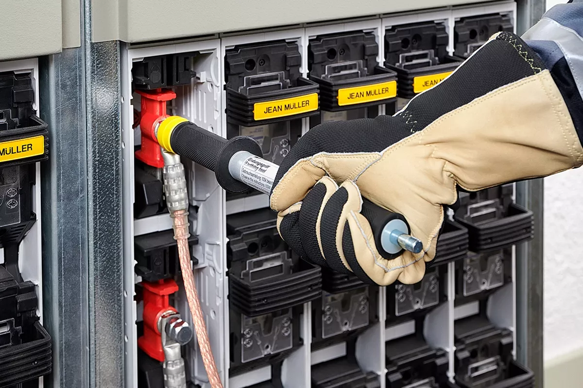 Close-up of a gloved technician connecting a grounding tool to a fuse switch in an electrical distribution cabinet with labeled modules.
